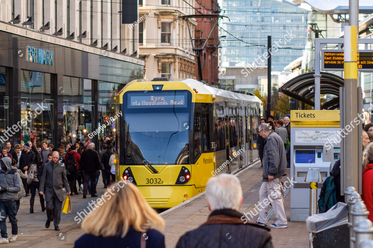 A single-lane tramway and station with a tram stopped at it in the centre of Manchester. The tramway is more or less part of the street, and there are people walking on and around it without difficulty, with shops lining the left-hand side of the street. The street is dominated by the people and pedestrian-space, with the tram fitting nicely into this. The station has level-boarding with almost no gap between it and the tram.